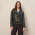 A woman modeling a black leather moto jacket with a zippered front, belted waist, and flap chest pocket, worn over a light pink collared shirt and dark trousers against a textured beige wall.