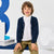 A young boy wearing a navy blue knit cardigan over a white collared shirt and olive green trousers, sitting against a white studio background with large blue and white decorative letters.