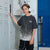 A child model wearing a dark gray t-shirt with a white speckled gradient pattern and 'NEW YORK' printed on the chest, standing in front of gray and blue lockers.