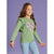 A young girl with long brown hair wearing a sage green long-sleeve top with a purple and white floral heart on the chest, paired with light blue jeans, standing against a solid purple background.