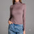 A woman wearing a fitted, long-sleeve turtleneck top in a muted dusty mauve color, paired with blue jeans, against a solid gray studio background.