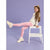 A young girl with long brown hair sits on a small white step stool, wearing pink polka-dot leggings, a cream sweatshirt, and cream sneakers against a solid lavender studio background.