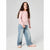 A young girl with long brown hair stands against a light gray studio background, wearing a light pink long-sleeve top with small black polka dots, light blue wide-leg jeans, and pale pink shoes.
