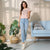 A woman modeling a light pink t-shirt with a sequined heart design, paired with light-wash blue skinny jeans and white sneakers, standing in a modern room with a light blue sofa and potted plants.