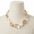 A statement necklace with gold-toned polished and faceted beads, white pearl-like beads, and clear glass beads, displayed on a beige fabric mannequin bust.