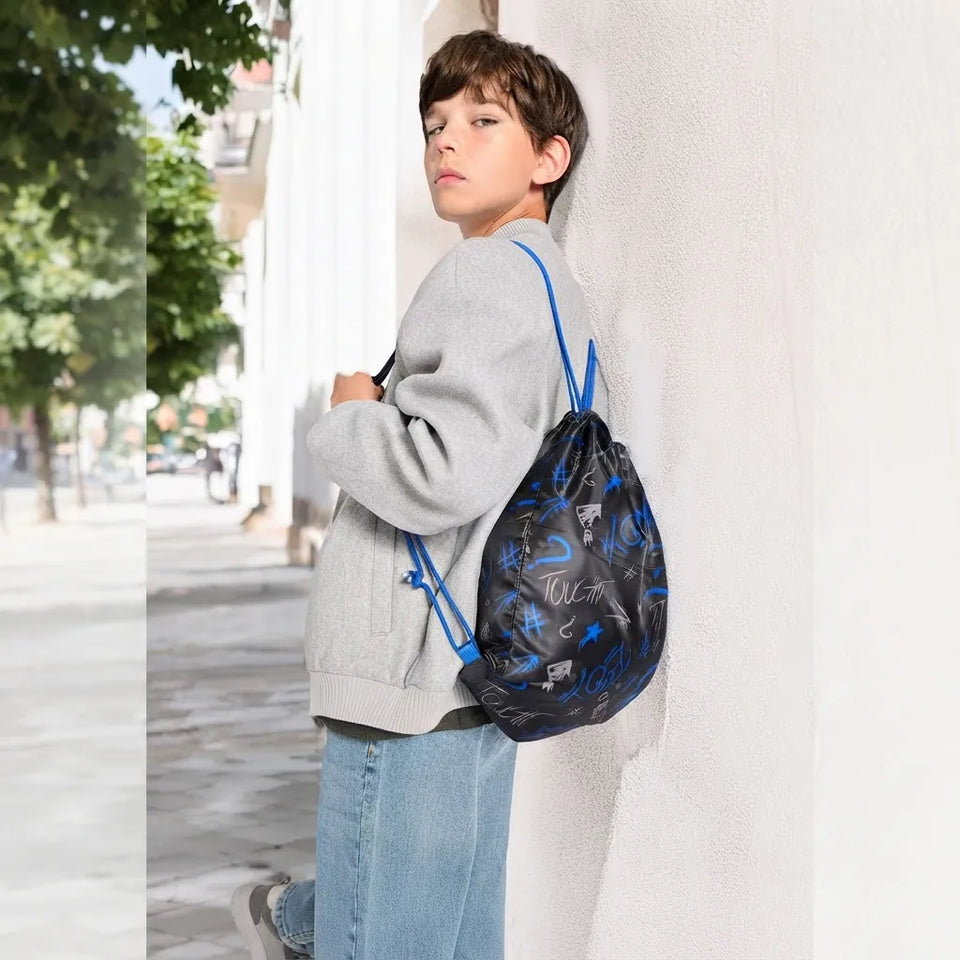 A child wearing a gray sweatshirt and blue jeans stands against a white wall, carrying a black drawstring backpack with blue graffiti-style lettering and blue straps.