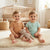 Two smiling toddlers sitting on a fluffy cream rug in a nursery, wearing matching short-sleeve henley outfits. The child on the left wears a warm tan/brown romper, and the child on the right wears a light mint green romper.