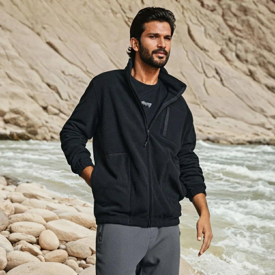 A man standing on a rocky riverbank wearing a black zip-up fleece jacket over a black t-shirt and gray sweatpants, with a sandy cliff and rushing water in the background.