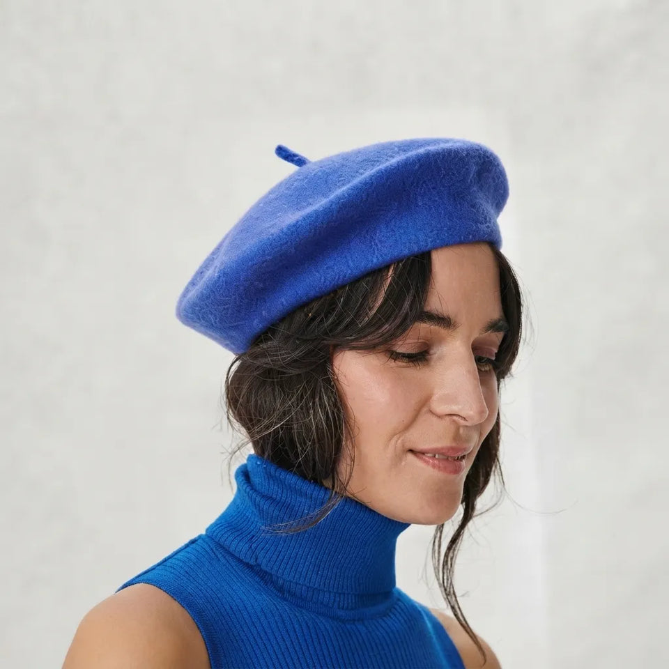 A woman wearing a bright royal blue felt beret and a matching blue ribbed turtleneck sweater, shown in a close-up portrait against a light textured wall.