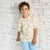 A young boy with curly brown hair stands against a white brick wall wearing a cream-colored short-sleeve t-shirt with a black line-art fish pattern and light blue pants.