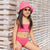 A young girl wearing a bright pink two-piece swimsuit with a matching pink textured bucket hat and pink sunglasses, posing in front of a white wooden slatted wall.