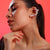 A close-up of a woman with her hair in a ponytail, wearing a curved cluster ear cuff made of white faux pearls and small red rhinestone accents, set against a solid coral-pink background.