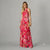 A woman modeling a sleeveless, high-neck, floor-length floral dress in vibrant pink and red tones with green leaves, set against a solid gray studio background.