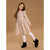 A young girl modeling a beige long-sleeve dress with black polka dots, white tights, and black boots, standing against a solid tan studio background.