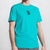A man wearing a bright turquoise crewneck t-shirt with a small black graphic of a mug on the left chest, shown against a light gray background.