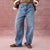 A person wearing light-wash wide-leg blue jeans, styled with a brown jacket featuring white shearling lining and brown suede shoes, standing against a textured pink backdrop.