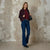 A woman stands against a textured plaster wall wearing a dark burgundy bomber jacket over a burgundy top, blue flare jeans, and black ankle boots, while holding a black handbag.