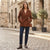 A woman stands on a cobblestone street wearing a brown belted wrap coat, dark blue jeans, and brown ankle boots, with a small brown handbag and necklace.