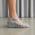A person wearing a light blue textured wedge sandal with crisscrossing straps and an ankle buckle, shown from the side against a neutral gray background.