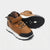 A pair of mid-top hiking boots in brown and black with white soles, shown on a light gray background. One boot is positioned upright to show the side profile, while the other lies on its side to display the textured black outsole.