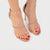 A pair of women's high-heeled sandals with metallic gold straps and rhinestone detailing, shown on a model's feet with red toenail polish against a light gray background.