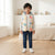 A young boy stands in a bright, modern bedroom wearing a light gray long-sleeve puffer vest with a colorful dinosaur print, blue jeans, and white sneakers.
