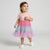 A toddler girl wearing a pink short-sleeve t-shirt with a unicorn graphic and a tiered tulle tutu skirt in pink, purple, and light blue, standing against a light gray studio background.