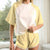 A woman wearing a two-piece lounge set featuring a short-sleeve raglan t-shirt and matching shorts in pale yellow and cream, with subtle yellow text graphics on the chest and thigh.