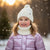 A young girl smiling outdoors in falling snow, wearing a white knit pom-pom beanie, a cream ribbed neck warmer, and a shiny lavender puffer jacket.
