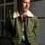 A man wearing an olive green bomber jacket with a cream sherpa collar, layered over a plaid shirt, standing against a dark textured wall.
