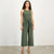 A woman modeling a sleeveless olive green jumpsuit with a matching waist tie, photographed against a plain light gray studio background.