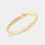 A gold bangle bracelet with a textured exterior, a small oval charm, and a row of tiny clear stones, shown on a light gray background.