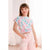 A young girl wearing a short-sleeve t-shirt with a pastel pink, blue, and white pattern of cartoon stars and hearts, paired with light pink pants, standing in a brightly lit room.