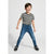 A young boy smiling and posing in a black and white striped short-sleeve t-shirt, blue skinny jeans, and black sneakers against a light gray studio background.