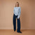 A woman stands against a warm beige studio backdrop wearing a light blue knit sweater and dark navy wide-leg trousers.