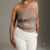 A woman wearing a strapless ruched tube top in taupe with a subtle blue-gray gradient and white pants, photographed against a neutral gray studio background.