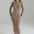 A woman wearing a fitted, long-sleeve ribbed knit midi dress in a taupe-brown color, shown from the neck down against a neutral gray studio background.