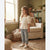 A young girl stands in a sunlit living room wearing a cream puff-sleeve smocked top, light-wash blue jeans, and white sneakers.