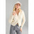 A woman modeling a cream ribbed knit zip-up hoodie with a half-zip collar, paired with gray jeans and a matching cream beanie, against a solid gray studio background.