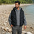 A man standing on a rocky riverside beach wearing a black puffer vest over a gray hoodie and gray pants.