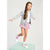 A young girl with long brown hair stands on a mint green pedestal, wearing a light-wash denim jacket, a white t-shirt, a pink floral mini skirt, and white sneakers with pink straps, against a light gray studio background.