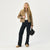 A blonde model stands against a light gray studio background wearing a cropped tan quilted jacket over a white top, dark wide-leg jeans, tan shoes, and holding a small black handbag.
