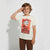 A young boy wearing a cream-colored t-shirt with a red and black graphic of Marshall the Dalmatian from Paw Patrol, paired with brown pants, saluting against a light gray background.