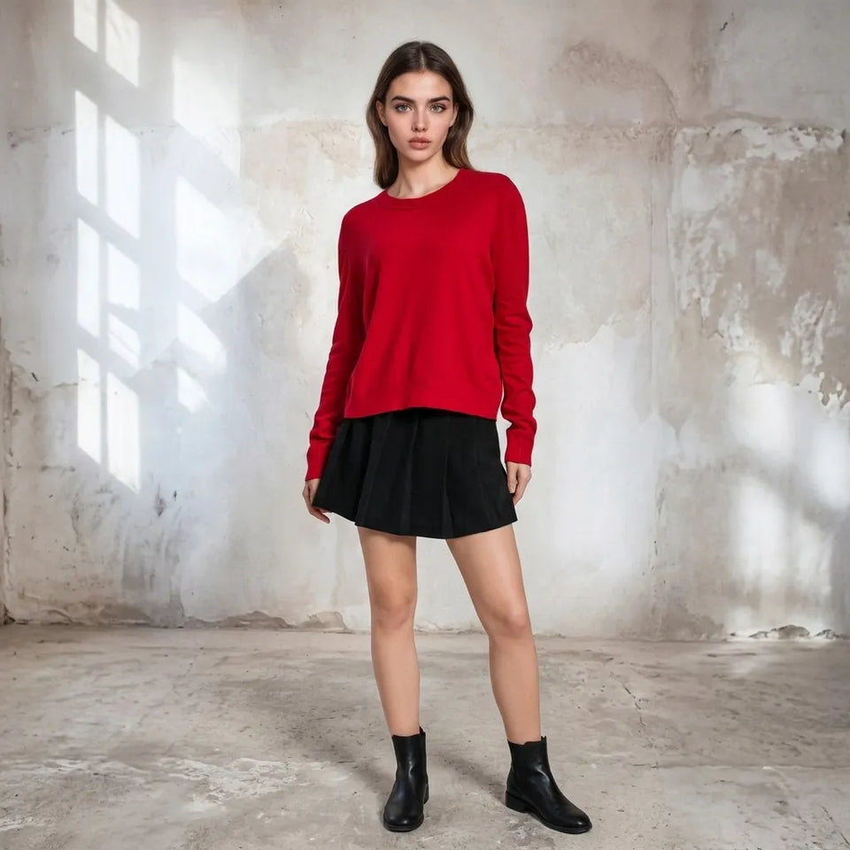 A woman stands in a rustic studio setting wearing a bright red long-sleeve knit sweater, a black pleated mini skirt, and black ankle boots.