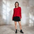 A woman stands in a rustic studio setting wearing a bright red long-sleeve knit sweater, a black pleated mini skirt, and black ankle boots.