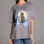 A woman wearing an oversized gray t-shirt with a graphic of a black cat sitting in front of a sun and clouds, paired with blue jeans, against a solid gray background.