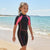 A young girl smiling on a sandy beach wearing a black and pink short-sleeve wetsuit, with the blue ocean and sky in the background.
