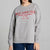 A woman wearing a light gray crewneck sweatshirt with red text that reads 'SELF LOVE CLUB' and smaller supportive phrases underneath, paired with blue jeans against a plain light gray studio background.