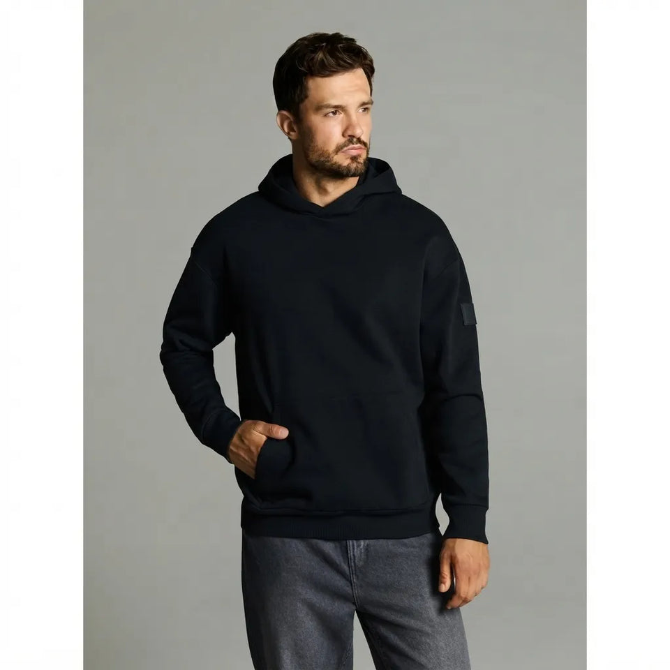 A man wearing a black pullover hoodie with a kangaroo pocket and ribbed cuffs, paired with gray jeans, standing against a solid gray studio background.