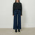 A model stands against a plain light gray studio wall wearing a black ribbed long-sleeve sweater, dark blue wide-leg denim jeans, and black ankle boots.